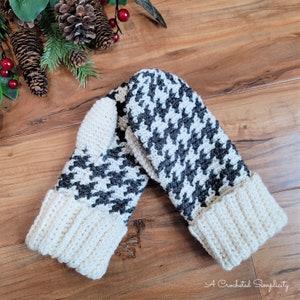 May include: A pair of crocheted mittens with a black and white houndstooth pattern and a cream-colored cuff. The mittens are lying on a wooden surface.