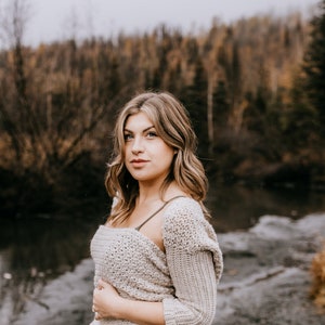 May include: A woman wearing a light brown crocheted sweater with a hood and a dark green skirt. She is standing in front of a river with trees in the background.