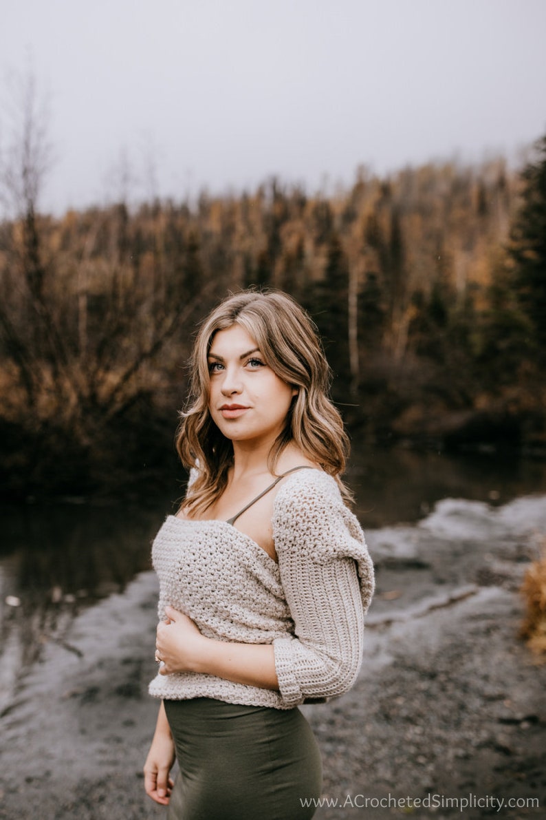 May include: A woman wearing a light brown crocheted sweater with a hood and a dark green skirt. She is standing in front of a river with trees in the background.