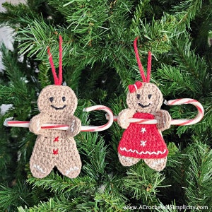 May include: Two crocheted gingerbread people ornaments hanging on a Christmas tree. The ornaments are holding candy canes. One gingerbread person is wearing a red dress with white snowflakes. The other gingerbread person is wearing a brown outfit.