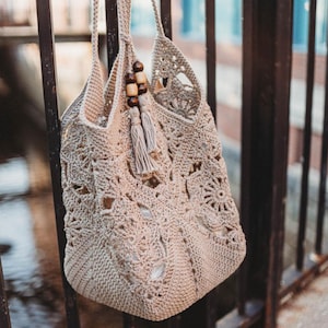 May include: A beige crochet tote bag with a floral and geometric pattern. The bag has two handles and is adorned with wooden beads and a tassel. The bag is hanging on a black metal fence.