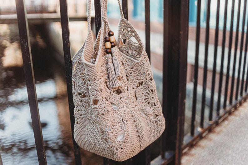 May include: A beige crochet tote bag with a floral and geometric pattern. The bag has two handles and is adorned with wooden beads and a tassel. The bag is hanging on a black metal fence.