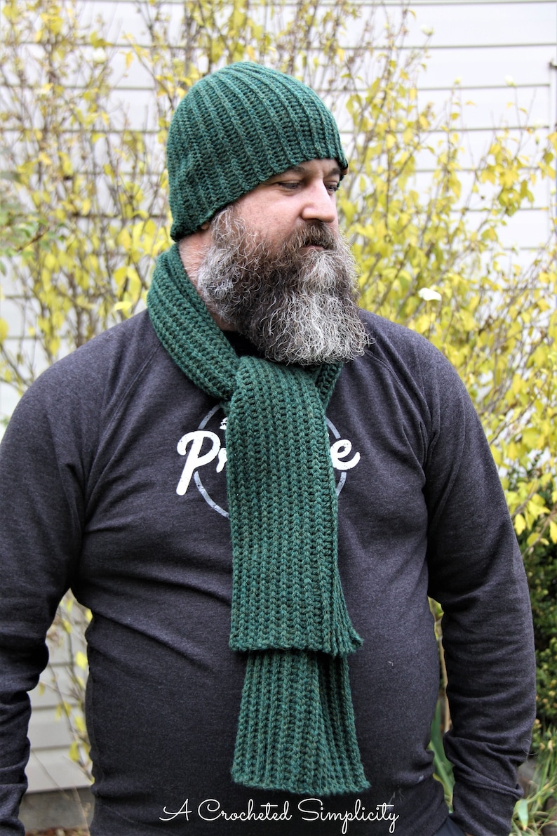 K&ouml;nnte beinhalten: Ein gr&uuml;nes H&auml;kelm&uuml;tzen- und Schals-Set. Die M&uuml;tze ist im Stil einer lockeren Beanie und der Schal ist lang und breit. Das Set wird von einer Person mit Bart getragen.