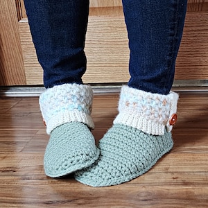 May include: A pair of crocheted slippers with a green base and a white and blue crocheted cuff. The slippers have a button on the top of the cuff.
