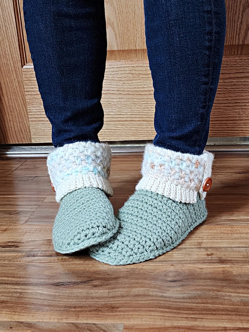 May include: A pair of crocheted slippers with a green base and a white and blue crocheted cuff. The slippers have a button on the top of the cuff.