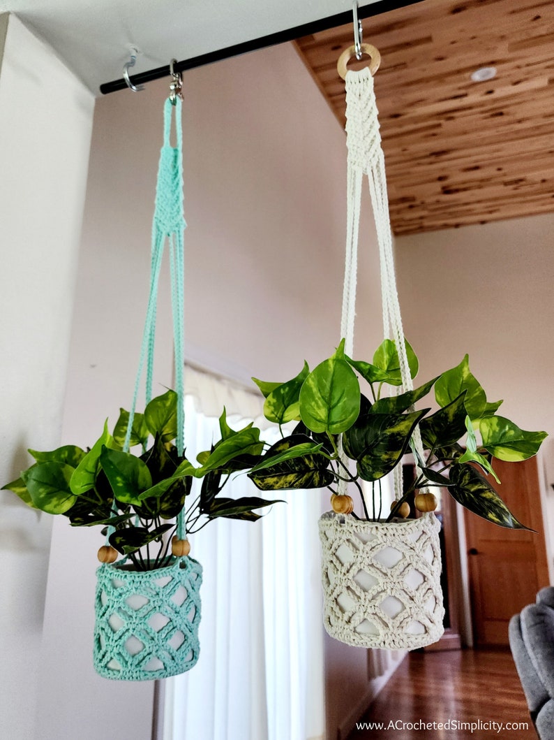 May include: Two macrame plant hangers with wooden rings, one is white and the other is light blue. Both hangers are holding potted plants with green leaves.