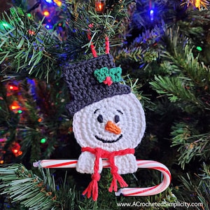 May include: A crocheted snowman ornament with a black hat, red scarf, and a candy cane. The snowman is smiling and has a carrot nose.