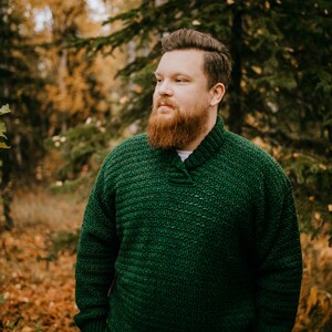 Pu&ograve; includere: Un maglione all'uncinetto verde scuro con collo a scialle. Il maglione &egrave; indossato da una persona in piedi all'aperto in un ambiente forestale. Il testo "A Crocheted Simplicity" &egrave; visibile nella parte inferiore dell'immagine.