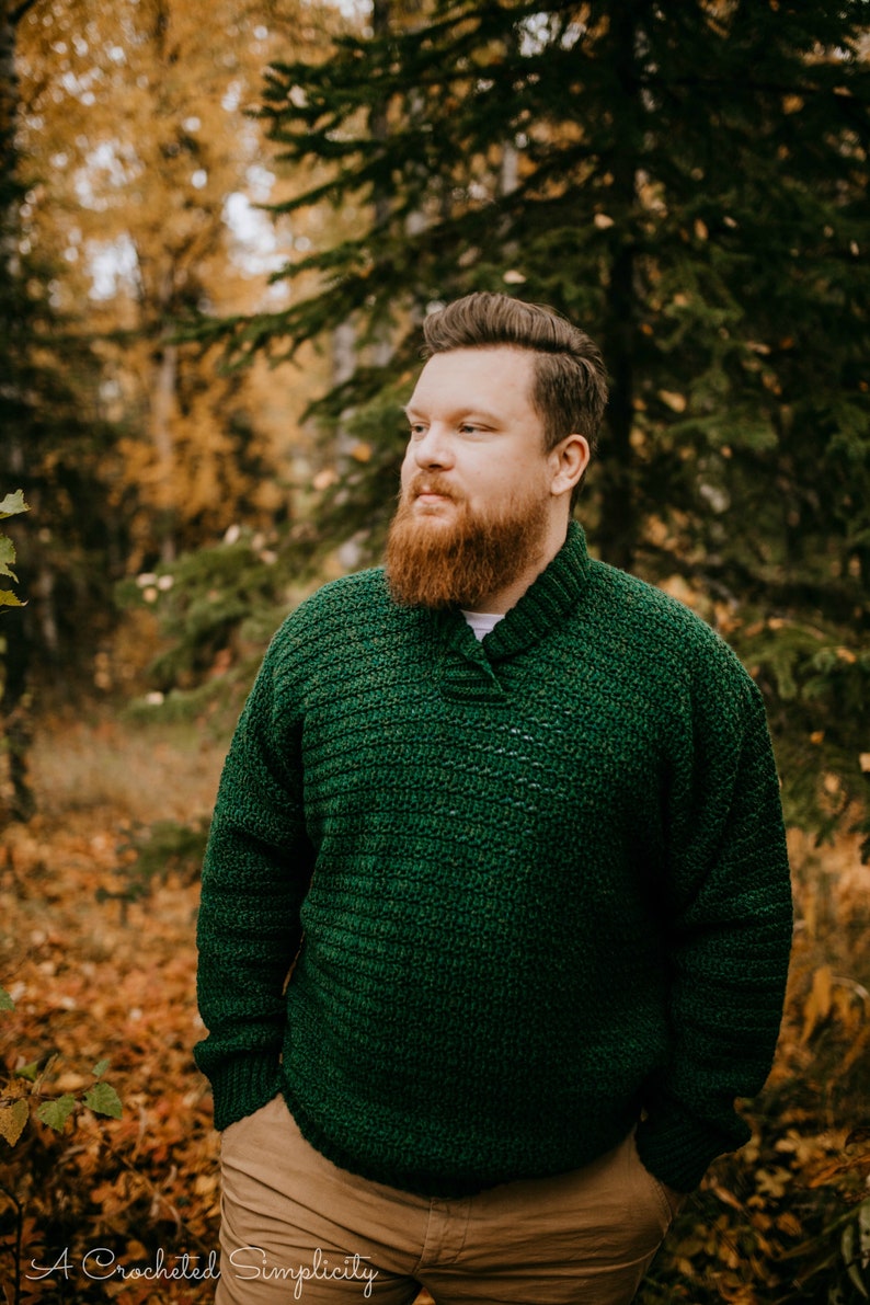 Pu&ograve; includere: Un maglione all'uncinetto verde scuro con collo a scialle. Il maglione &egrave; indossato da una persona in piedi all'aperto in un ambiente forestale. Il testo "A Crocheted Simplicity" &egrave; visibile nella parte inferiore dell'immagine.