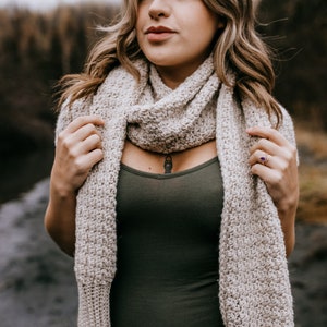 May include: A woman wearing a beige crocheted scarf. The scarf is draped around her neck and shoulders. She is wearing a dark green tank top.