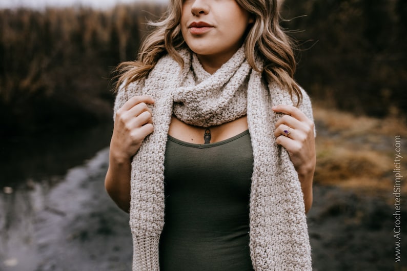 May include: A woman wearing a beige crocheted scarf. The scarf is draped around her neck and shoulders. She is wearing a dark green tank top.
