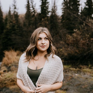 May include: A woman wearing a white crocheted cardigan with a green tank top underneath. She is standing in a forest setting with trees in the background.