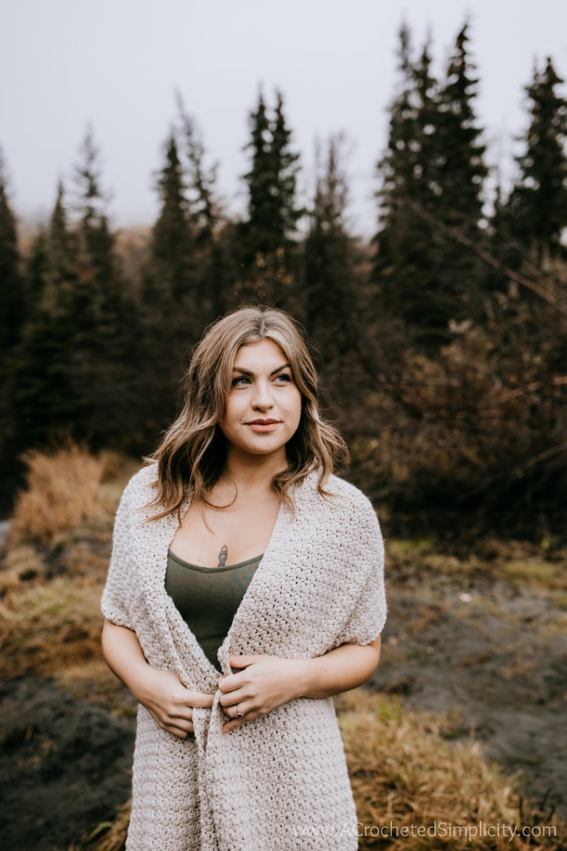 May include: A woman wearing a white crocheted cardigan with a green tank top underneath. She is standing in a forest setting with trees in the background.