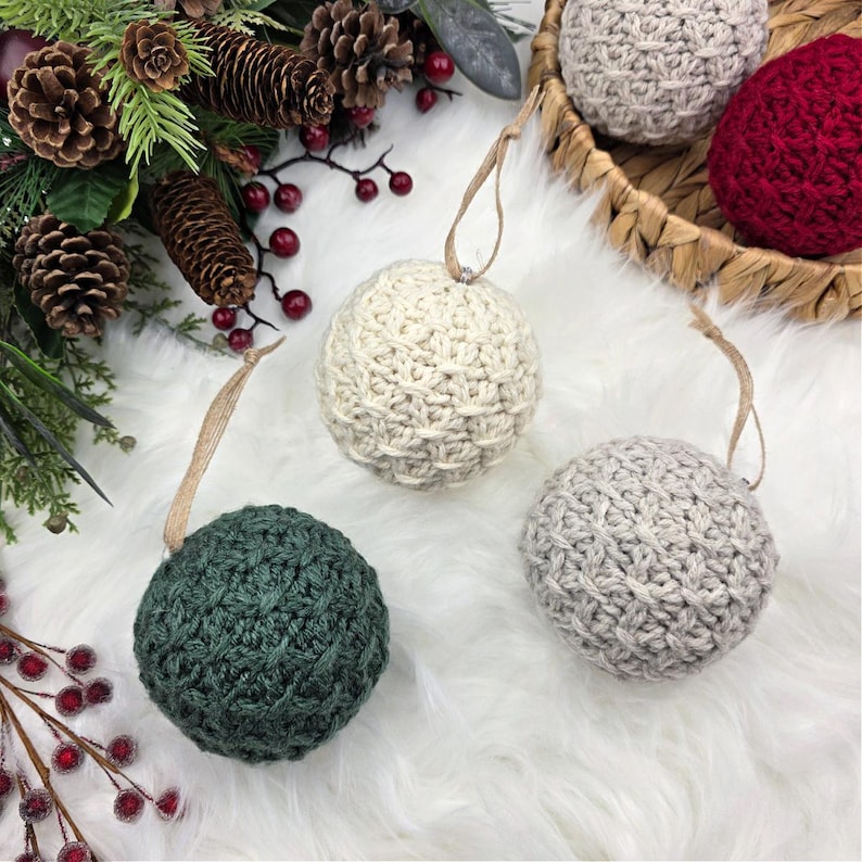 May include: A selection of handmade, crocheted Christmas baubles in various colours. The baubles are spherical, with a textured stitch pattern and a loop for hanging. Colours include cream, grey, green, and red. The baubles are displayed on a white, fluffy surface with pinecones and greenery.