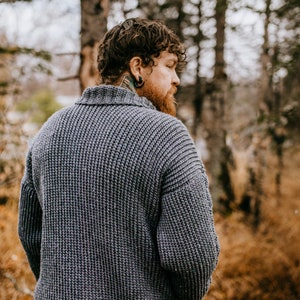 May include: A man wearing a gray knitted sweater with a ribbed texture, standing in a forest setting. The sweater has a high collar and long sleeves. The man is looking over his shoulder.