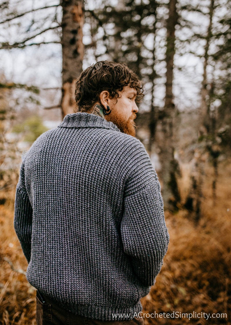 May include: A man wearing a gray knitted sweater with a ribbed texture, standing in a forest setting. The sweater has a high collar and long sleeves. The man is looking over his shoulder.