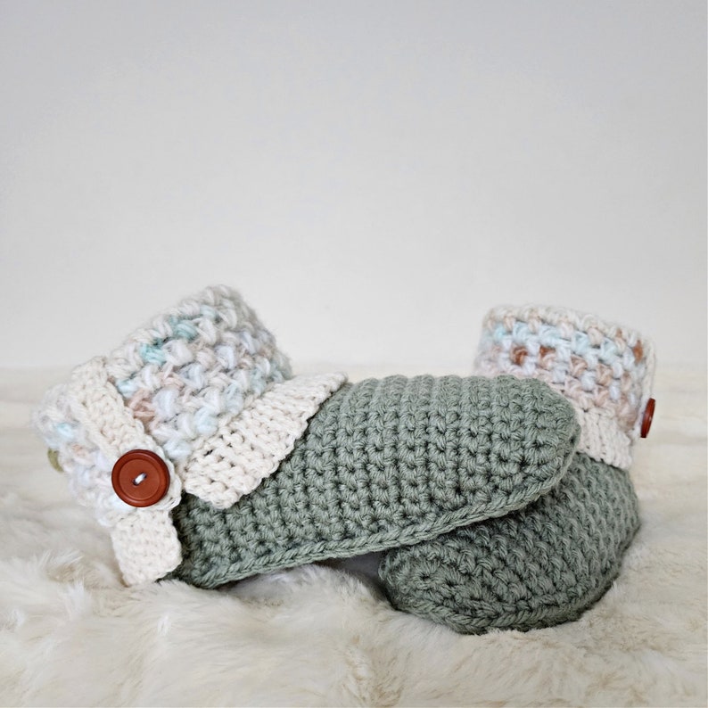 May include: A pair of crocheted slippers in a sage green color with a white and pastel colored crocheted cuff. One slipper has a brown button.