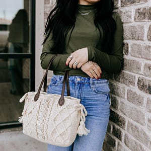 May include: Cream-colored, hand-knitted tote bag with brown leather handles. The bag features a cable knit design and diamond pattern. The bag has a drawstring closure with tassels. The woman is wearing a green long-sleeve shirt and blue jeans.