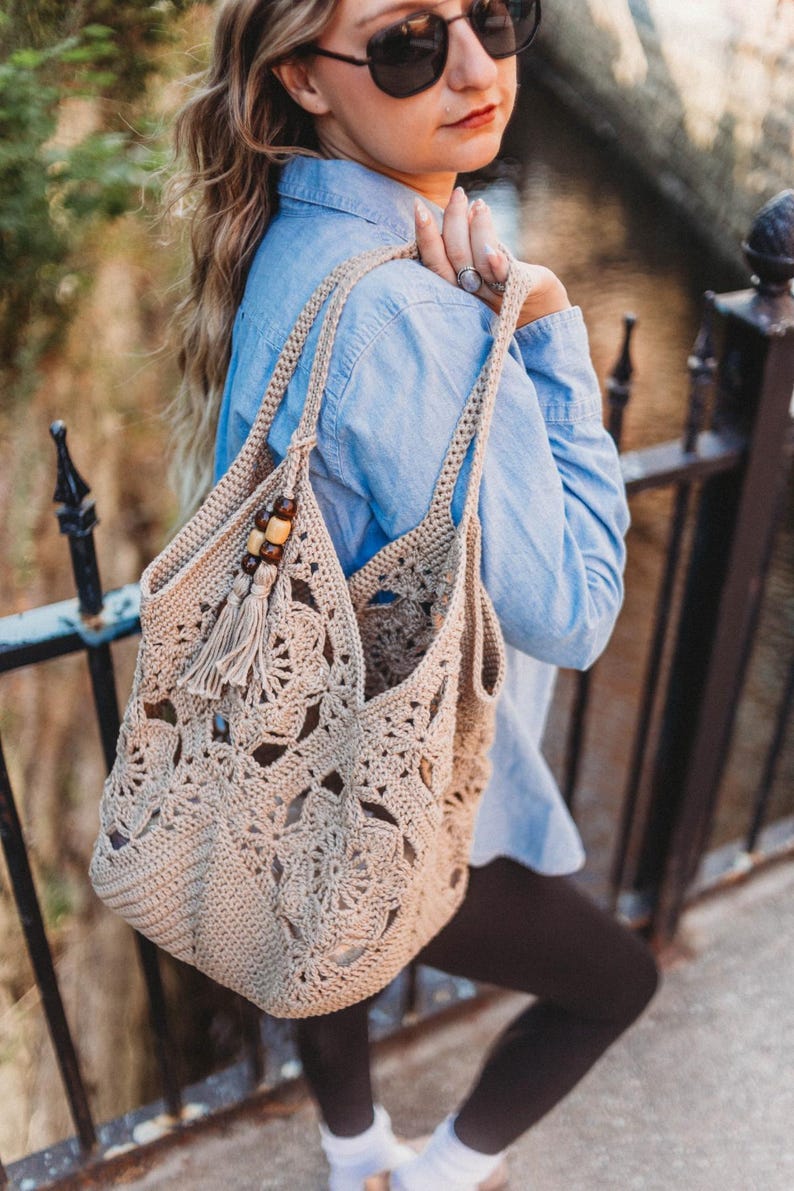 May include: A beige crochet tote bag with a floral pattern and a tassel. The bag has two handles and wooden beads. The bag is carried over the shoulder.