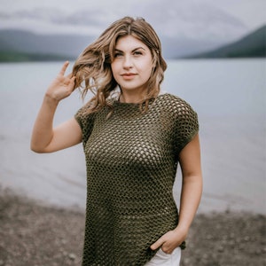 May include: A woman wearing a green crochet top with a loose, open weave. She is standing on a beach with a body of water in the background.