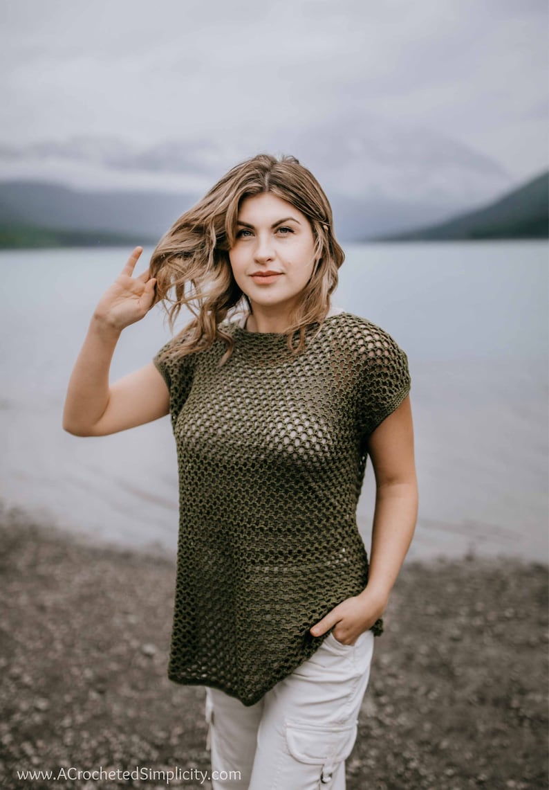 May include: A woman wearing a green crochet top with a loose, open weave. She is standing on a beach with a body of water in the background.
