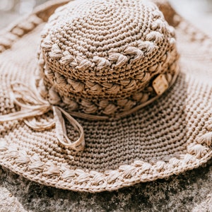 May include: A beige, crocheted sun hat with a wide brim and a decorative band around the crown. The hat features a bow and a small wooden tag. The hat is resting on a stone surface.