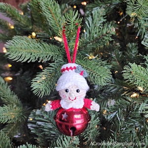 May include: Crocheted Christmas ornament shaped like a person wearing a white hat with a red trim and a red and white dress. The ornament is attached to a red jingle bell.