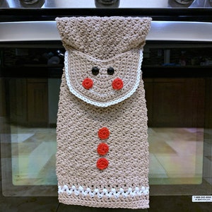 May include: A crocheted gingerbread man oven mitt with red buttons and a white trim. The oven mitt is hanging on a black oven door.
