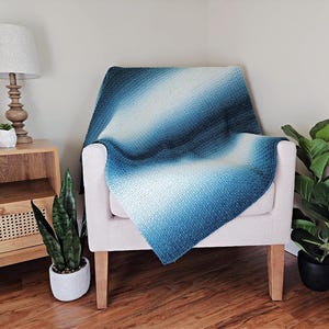 May include: A blue and white ombre knitted throw blanket draped over a white armchair. The blanket has a textured pattern and a gradient effect, transitioning from dark blue to light blue and white. The room is decorated with plants and a wooden side table.