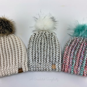Puede incluir: Tres gorros de punto con pompones de piel sintética. Los gorros son de color blanco, gris y rosa y turquesa. Cada gorro tiene una pequeña etiqueta de cuero marrón con el texto "A Crocheted Simplicity".