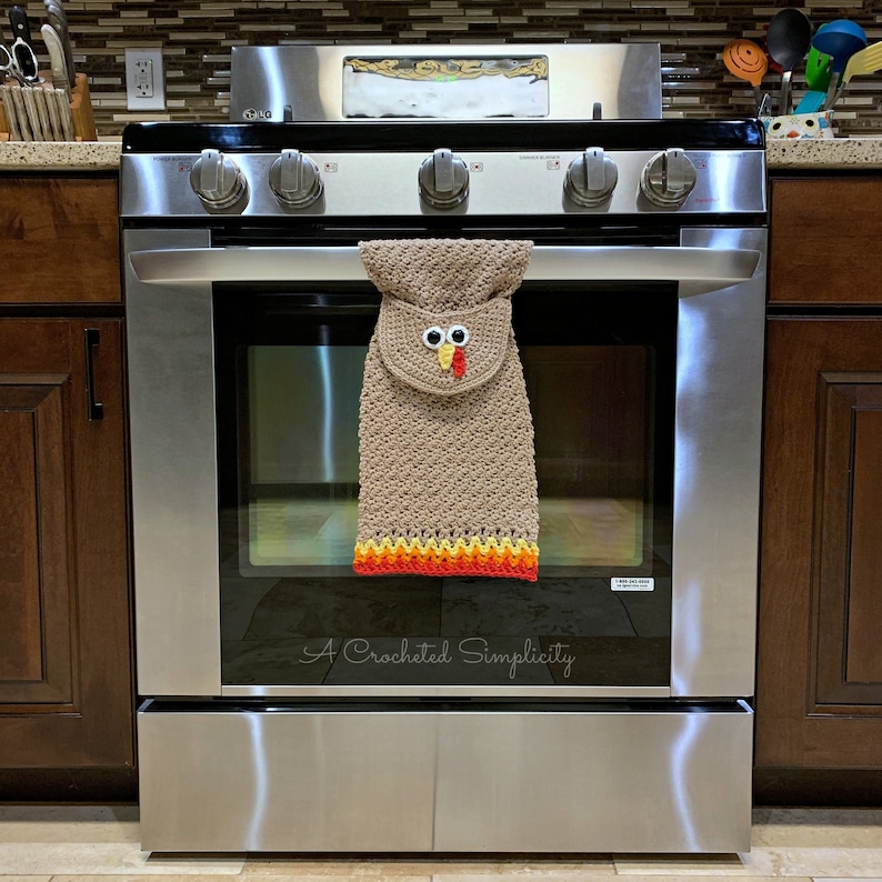 May include: A crocheted turkey oven mitt hanging on a stainless steel oven. The oven mitt is brown with orange, yellow, and red trim. The turkey has a white face with black eyes and a red beak. The text "A Crocheted Simplicity" is visible below the oven mitt.