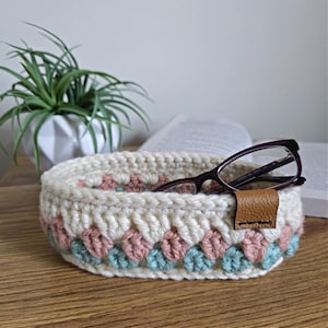 May include: A crocheted white oval-shaped bowl with a pink and teal trim. The bowl is filled with a pair of eyeglasses and has a brown leather strap.