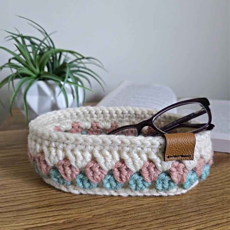 May include: A crocheted white oval-shaped bowl with a pink and teal trim. The bowl is filled with a pair of eyeglasses and has a brown leather strap.