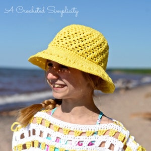 May include: A crocheted yellow sun hat with a wide brim. The hat is made of a textured yarn and is worn by a person. The image also shows a white and yellow crocheted top and the text "A Crocheted Simplicity".