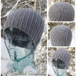 K&ouml;nnte beinhalten: Graue H&auml;kelm&uuml;tze auf einem Glas-Mannequin-Kopf. Die M&uuml;tze ist gerippt und hat ein schlichtes Design. Die M&uuml;tze ist vor einem verschneiten Hintergrund fotografiert.