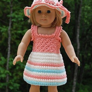 May include: A doll wearing a pink and white striped crocheted dress with a matching crocheted hat. The doll has blonde hair and blue eyes.