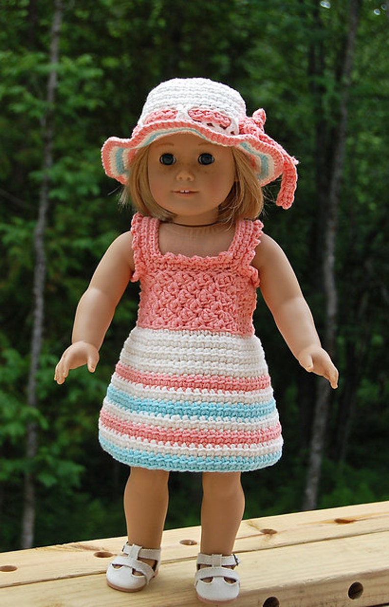 May include: A doll wearing a pink and white striped crocheted dress with a matching crocheted hat. The doll has blonde hair and blue eyes.
