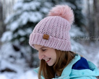 Può includere: Un berretto di lana rosa con un pompon soffice in cima. Il berretto ha un'etichetta in pelle marrone con il testo "A Crocheted Simplicity".