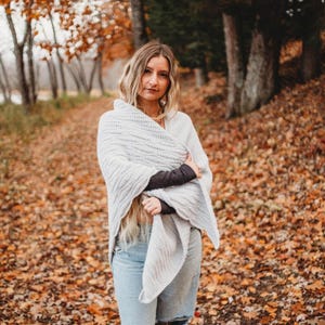 May include: A woman wearing a light gray shawl, wrapped around her shoulders, stands on a path covered in fallen autumn leaves. The shawl has a textured pattern. She is wearing a white top, dark long sleeves, and blue jeans.