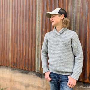 May include: A gray crocheted sweater with a collared neckline. The sweater has a ribbed texture and long sleeves. The model is wearing blue jeans and a gray baseball cap.