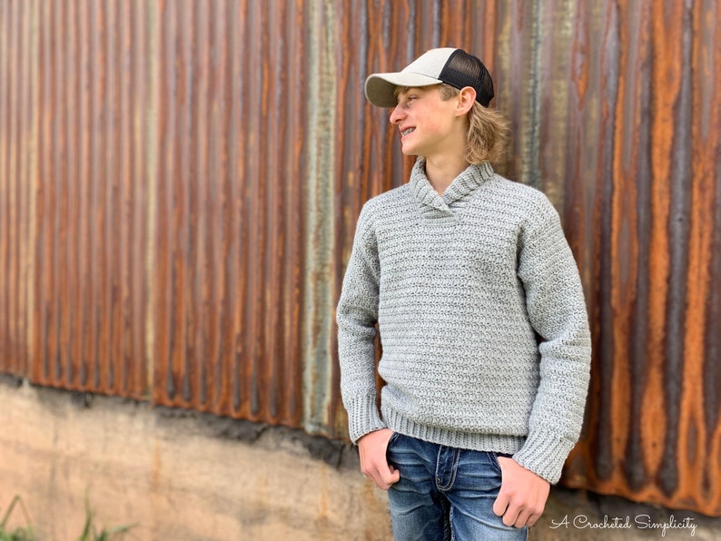 May include: A gray crocheted sweater with a collared neckline. The sweater has a ribbed texture and long sleeves. The model is wearing blue jeans and a gray baseball cap.