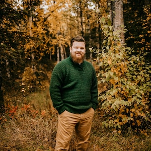 Pu&ograve; includere: Una persona indossa un maglione verde lavorato a maglia e pantaloni color cachi in una foresta con fogliame autunnale. Il maglione ha un motivo strutturato. Il testo "A Crocheted Simplicity" &egrave; in basso nell'immagine.