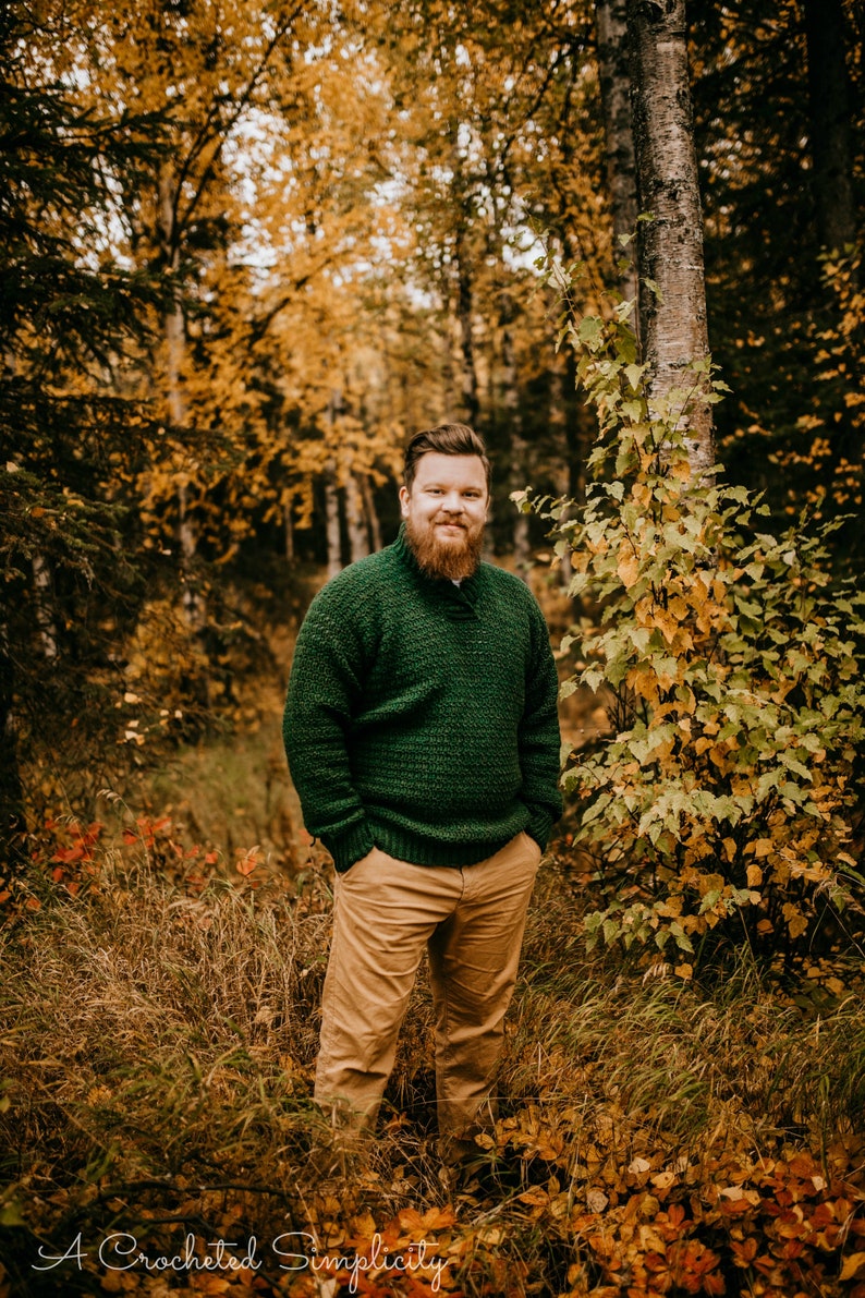 Pu&ograve; includere: Una persona indossa un maglione verde lavorato a maglia e pantaloni color cachi in una foresta con fogliame autunnale. Il maglione ha un motivo strutturato. Il testo "A Crocheted Simplicity" &egrave; in basso nell'immagine.