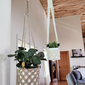May include: Two macrame plant hangers with wooden rings, one beige and one white, hanging from a black metal rod. The beige hanger holds a potted plant with green leaves, while the white hanger holds a potted plant with green and yellow leaves.