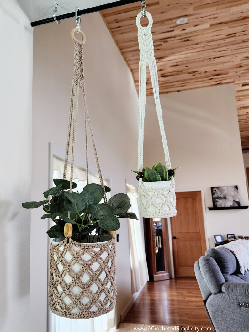 May include: Two macrame plant hangers with wooden rings, one beige and one white, hanging from a black metal rod. The beige hanger holds a potted plant with green leaves, while the white hanger holds a potted plant with green and yellow leaves.