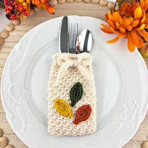 May include: A cream-colored, knitted cutlery holder with a drawstring closure, holding a knife, fork, and spoon. The holder is decorated with three crocheted leaves in yellow, green, and orange. The holder is placed on a white plate.