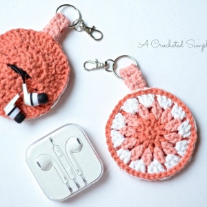 May include: Two crocheted headphone holders with keychains. The holders are round and made with pink and white yarn. The text "A Crocheted Simplicity" is visible in the top right corner of the image.
