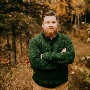 Pu&ograve; includere: Una persona indossa un maglione all'uncinetto verde scuro e strutturato con colletto abbottonato, abbinato a pantaloni color cachi. Lo sfondo presenta una foresta con fogliame autunnale, creando un ambiente caldo e naturale.
