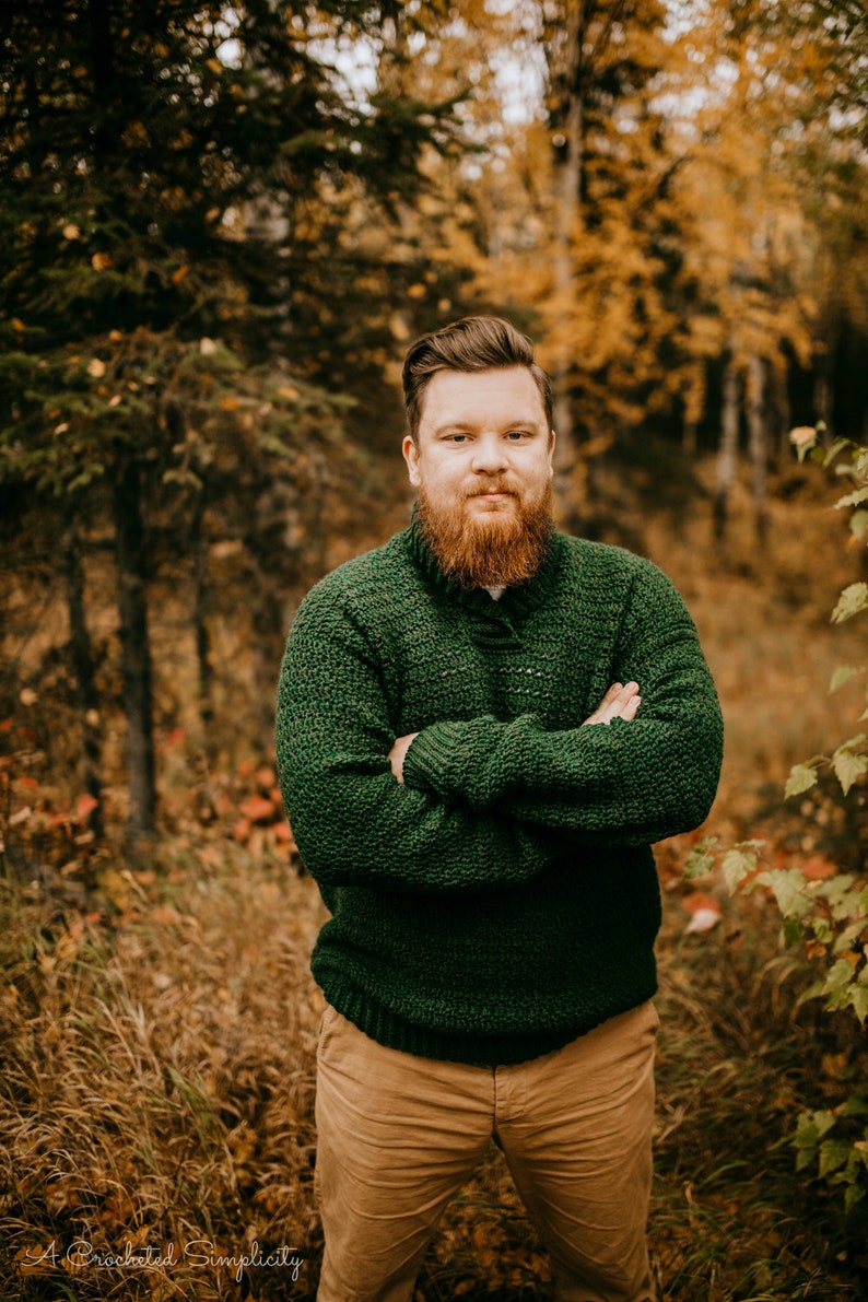 Pu&ograve; includere: Una persona indossa un maglione all'uncinetto verde scuro e strutturato con colletto abbottonato, abbinato a pantaloni color cachi. Lo sfondo presenta una foresta con fogliame autunnale, creando un ambiente caldo e naturale.