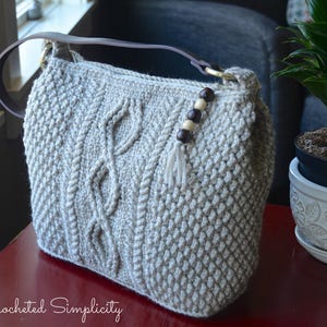 May include: A crocheted tote bag with a cable knit pattern and brown wooden beads. The bag is light gray and has a brown leather strap.